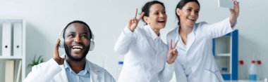 Kulaklıklı gülümseyen Afrikalı Amerikalı doktor klinikte akıllı telefondan selfie çeken bulanık meslektaşlarının yanında. 