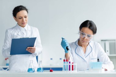 Scientist in goggles holding electronic pipette near african american colleague with clipboard and test tubes  clipart