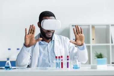 African american scientist in vr headset sitting near blurred medical equipment clipart