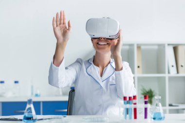 VR kulaklıklı gülümseyen bilim adamı bulanık ön planda test tüplerinin yanında oturuyor. 