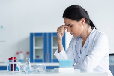 Şişelerin ve test tüplerinin yanında oturan yorgun bilim adamının yan görüntüsü 