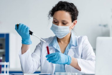Tıbbi maskeli Afrikalı Amerikalı bilim adamı ve plastik eldivenli elektronik pipet ve test tüpünü bulanık dizüstü bilgisayarın yanında tutuyor. 