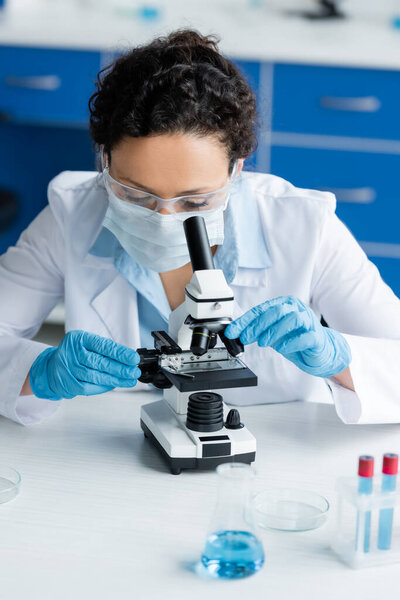 African american scientist in safety goggles and medical mask working with microscope near test tubes 