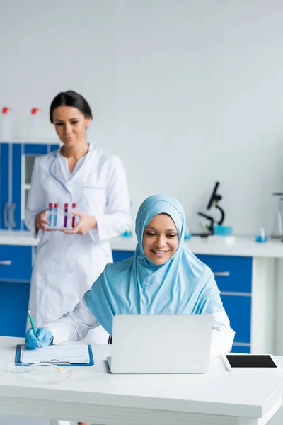 Smiling Muslim Scientist Using Laptop Writing Clipboard Blurred Colleague — Stock fotografie