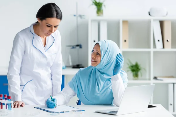 穿着乳胶手套的快乐的穆斯林科学家在剪贴板上写字 并在实验室里看着笔记本电脑旁边的同事 — 图库照片