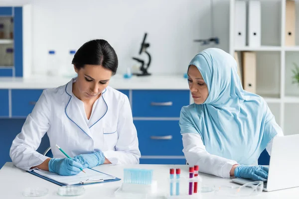 Scientist Latex Gloves Writing Clipboard Arabian Colleague Using Laptop Laboratory — Stock Fotó