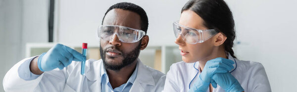 Multiethnic scientists in latex gloves and goggles looking at test tube in lab, banner 
