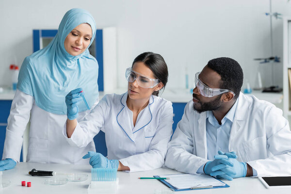Scientist holding test tube near interracial colleagues in safety goggles, electronic pipette and digital tablet 