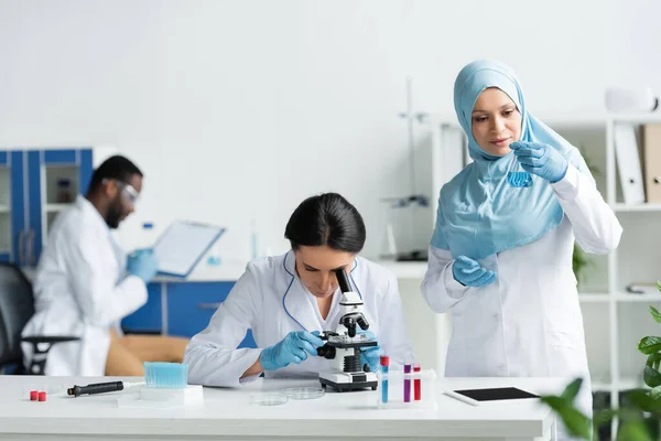 Cientista Árabe Segurando Frasco Com Reagente Perto Colega Com Microscópio — Fotografia de Stock