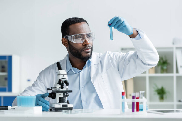 African american scientist in latex gloves and safety goggles holding test tube near microscope 