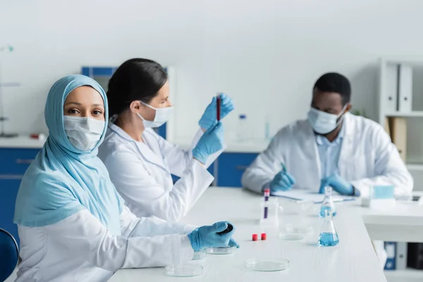 Muslim Scientist Medical Mask Holding Pipette Petri Dishes Blurred Colleagues — Stock fotografie