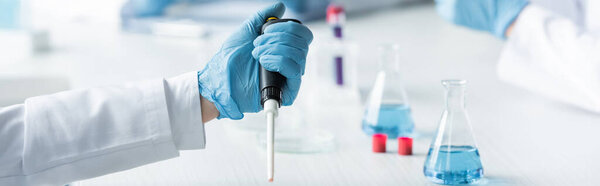 Cropped view of scientist in latex glove holding electronic pipette near flasks, banner 