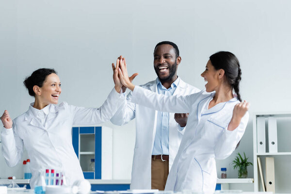 Cheerful multiethnic scientists giving high five near medical equipment in lab 