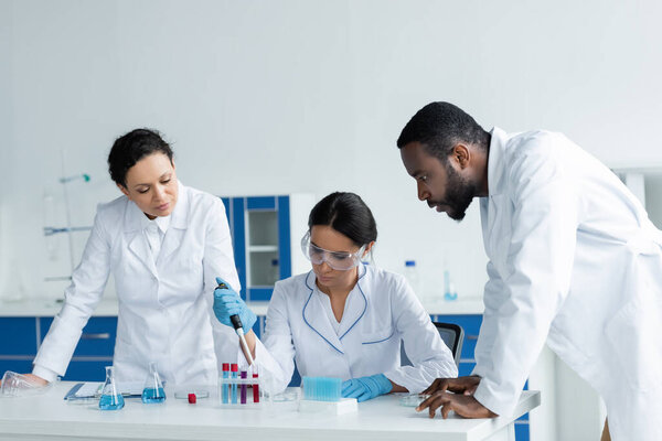 Scientist working with pipette and test tubes near focused colleagues in lab 