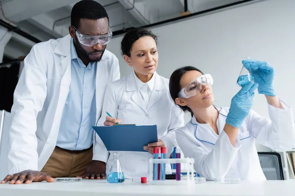 Interracial Scientists Safety Goggles Clipboard Looking Reagent Flask — Stock Photo, Image
