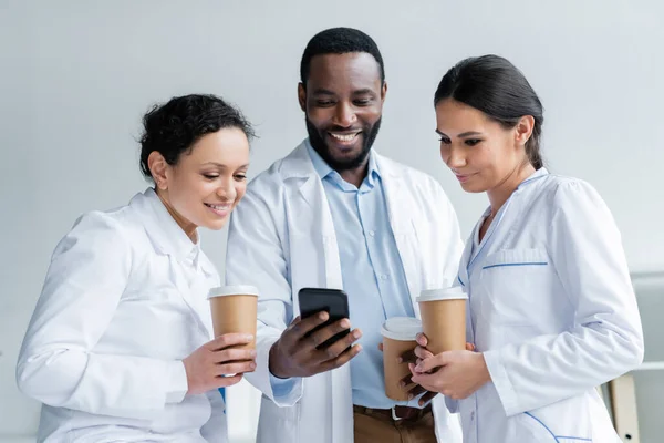 Αφροαμερικανός Γιατρός Κρατώντας Smartphone Κοντά Συναδέλφους Καφέ Στην Κλινική — Φωτογραφία Αρχείου