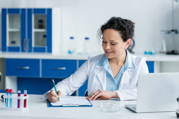 Glimlachend Afrikaans Amerikaans Wetenschapper Schrijven Klembord Buurt Van Laptop Petrischaaltjes — Stockfoto