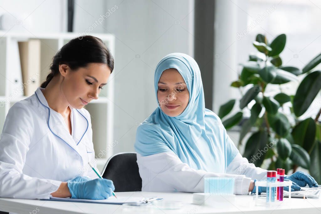 Muslim scientist looking at colleague writing on clipboard near test tubes on blurred foreground 