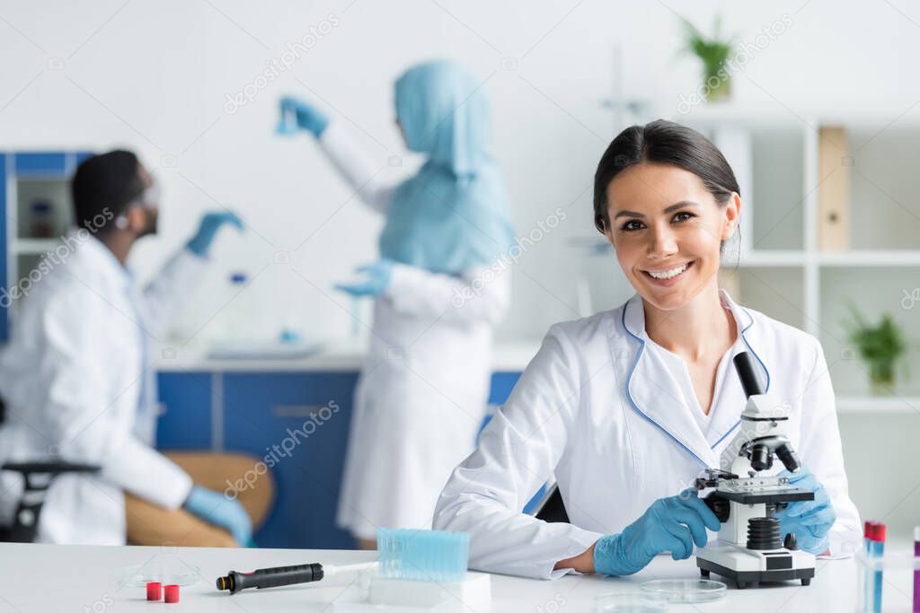 Happy scientist in latex gloves smiling at camera near microscope and test tubes 