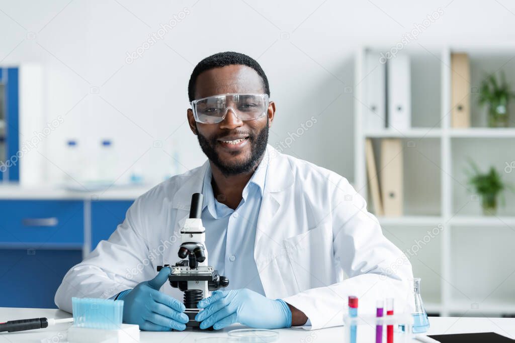 Smiling african american scientist in protective goggles looking at camera near microscope in lab 