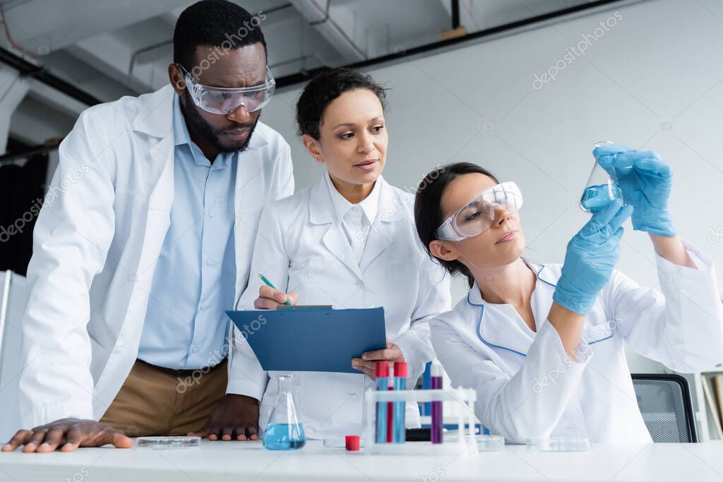Interracial scientists in safety goggles with clipboard looking at reagent in flask 