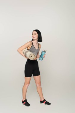 Vitiligo 'su olan neşeli sporcu kadın gri arka planda spor şişesi ve spor paspası tutuyor.