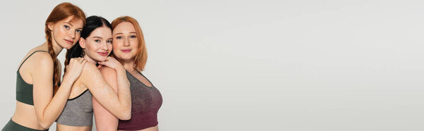 Body positive sportswomen hugging and smiling isolated on grey, banner 