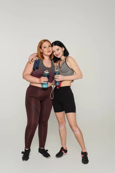 Sportswoman with vitiligo hugging friend with sports bottle and jump rope on grey background