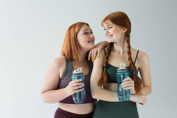 Plus size sportswoman with sports bottle looking at smiling red haired friend isolated on grey 