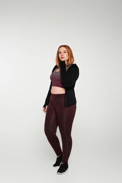 Red haired plus size woman in sportswear standing on grey background