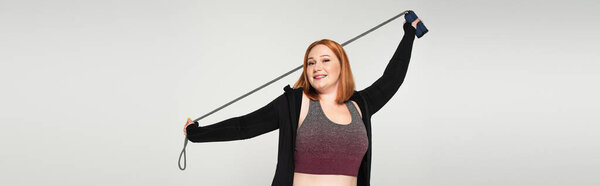 Red haired sportswoman holding jump rope isolated on grey, banner 