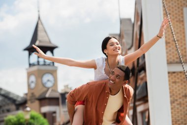 Neşeli Asyalı kadın dışarıda mutlu Afro-Amerikan erkeğini sırtında taşıyor.