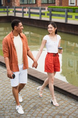 stylish multiethnic couple with paper cups holding hands while walking near water in city clipart