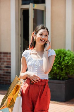 Alışveriş çantalı neşeli Asyalı kadın dışarıda cep telefonuyla konuşuyor.