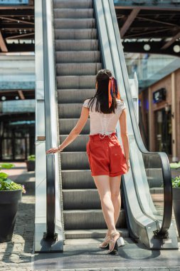 back view of young woman in shorts carrying shopping bags near escalator clipart