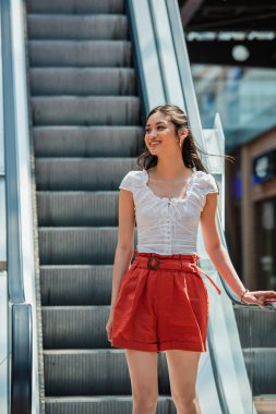 happy asian woman in summer clothes looking away on escalator clipart