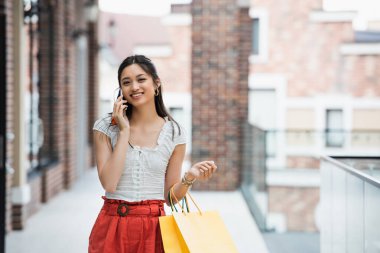 Alışveriş çantalı memnun Asyalı kadın şehir caddesinde cep telefonuyla konuşuyor.