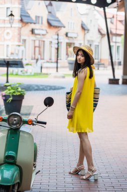 Sarı elbiseli ve güneş şapkalı güzel Asyalı kadın scooter 'ın yanındaki kameraya bakıyor.