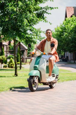 Yeşil caddede scooter sürerken kameraya bakan Afro-Amerikan bir adam