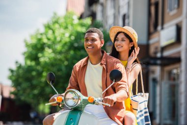 Afrikalı Amerikalı erkek arkadaşıyla scooter sürerken Hasır Şapka 'ya dokunan neşeli Asyalı kadın
