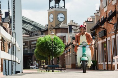 Şehrin caddesinde scooter süren koruyucu kasklı neşeli Afrikalı Amerikalı adam.