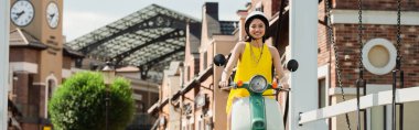 Kasketli mutlu Asyalı kadın şehir sokağında scooter sürmekten hoşlanıyor.