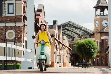 Sarı elbiseli ve kasklı genç Asyalı kadın şehir caddesinde scooter kullanıyor.