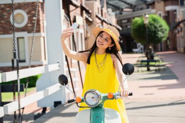 Scooterlı mutlu Asyalı kadın köprüde selfie çekiyor.