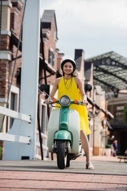Sarı elbiseli mutlu Asyalı kadın şehir sokağında scooter kullanıyor.