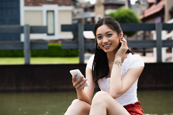 Весела Азіатська Жінка Мобільним Телефоном Дивиться Камеру Сидячи Відкритому Повітрі — стокове фото
