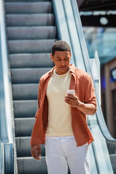身穿衬衫的年轻的非洲裔美国人在自动扶梯上看着智能手机 — 图库照片