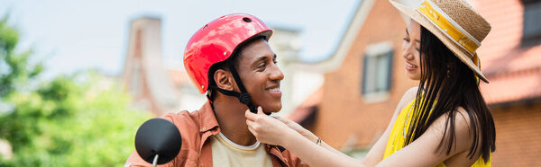 young asian woman in straw hat fastening hardhat of african american man, banner