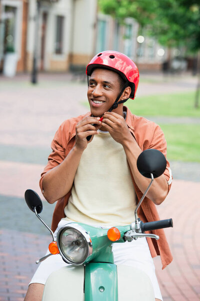 smiling african american man on scooter fastening protective helmet