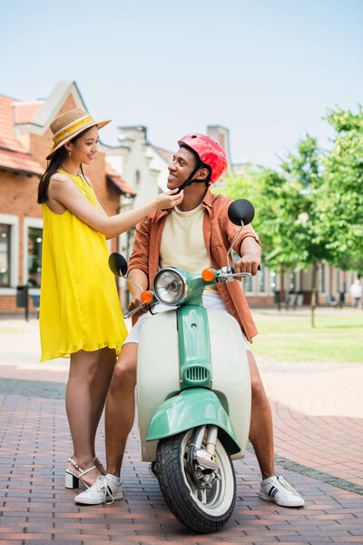 young asian woman in stylish clothes fastening helmet of african american man on scooter